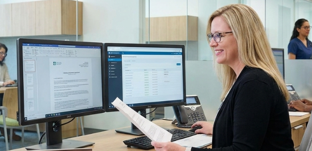 Smiling credentialing specialist holding a printed document at a desk with two monitors showing a letter and a status list
